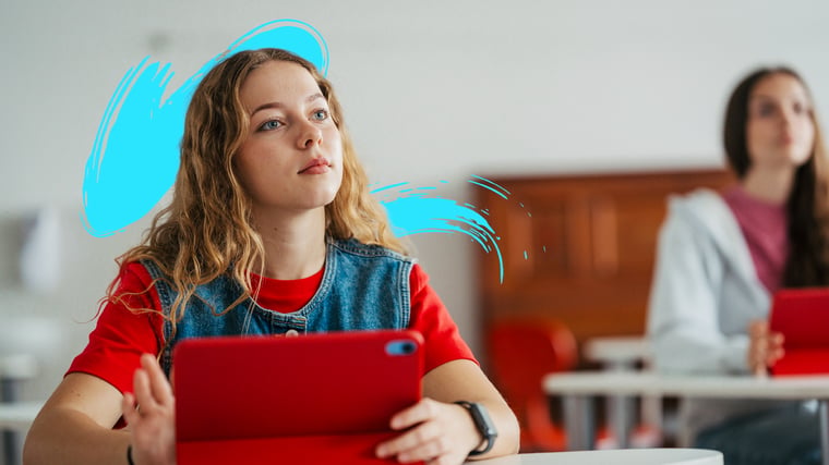 Two students sit in a classroom, one holding a red tablet and looking forward attentively.
