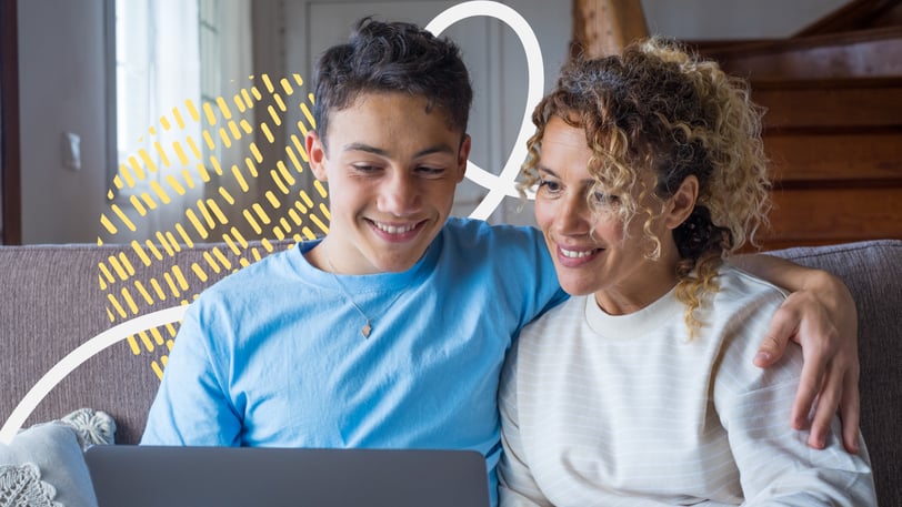  A teenager and his mother smile together while looking at a laptop on the couch at home.